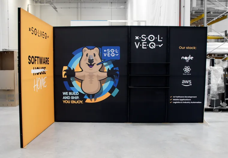 Stoisko od frontu: czarna ściana z maskotką quokką, logo Solveq, półkami i listą technologii
